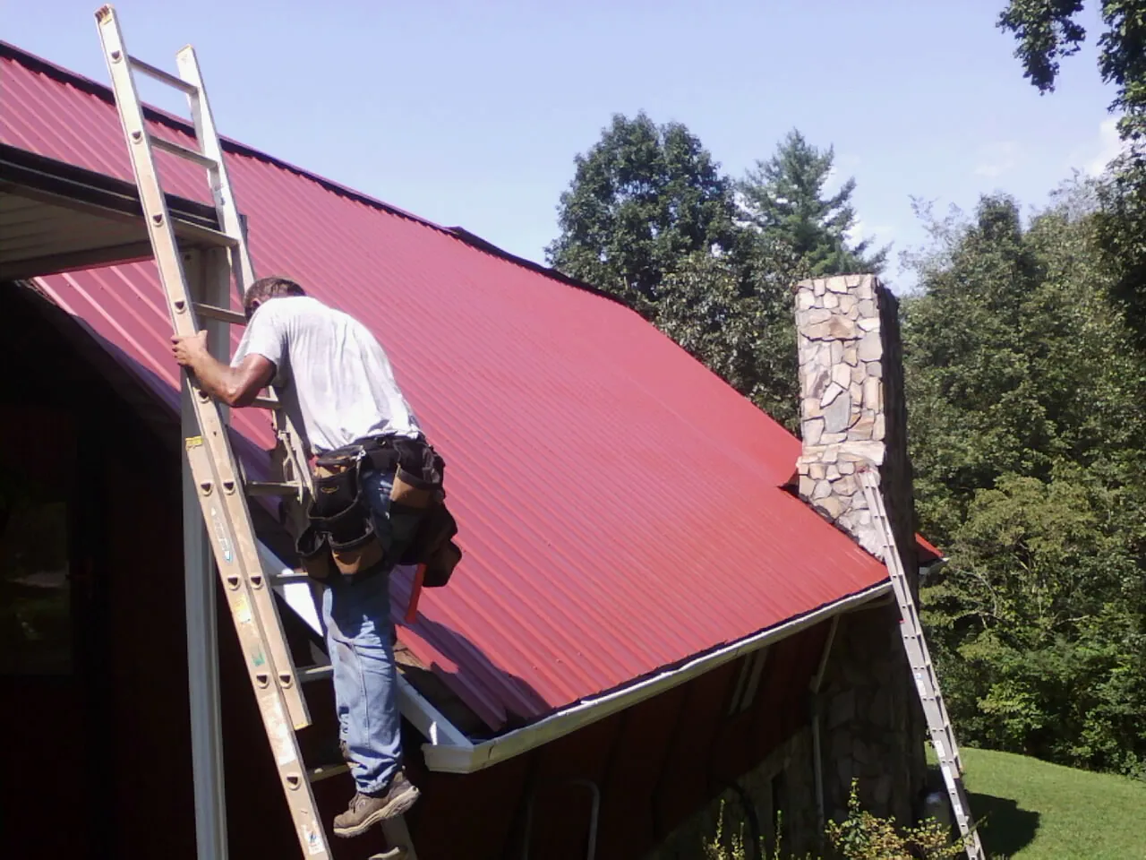 Roofer on a ladder beside a red metal roof and stone chimney — Soffit & Fascia Repair by Roofing Brothers in Plattsburgh